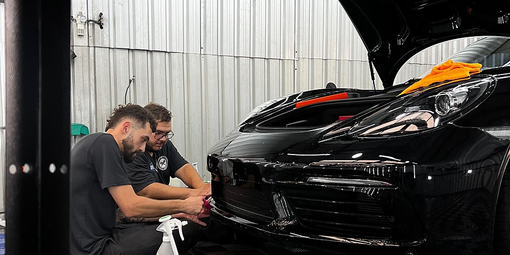 Dois profissionais da Lasmar Car Detail aplicando PPF na dianteira de um Porsche preto, com microfibra laranja sobre o capô aberto